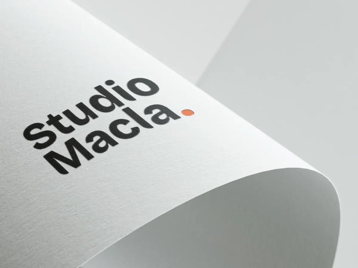 Logo Studio Macla imprimé en noir sur un papier texturé qui se courbe, le point en orange